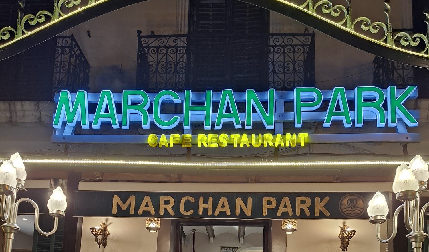 La façade de Marchan Park le soir : l'enseigne néon verte « Marchan Park Café Restaurant » sous le balcon en fer forgé, éclairée par les lampes-globes de l'entrée