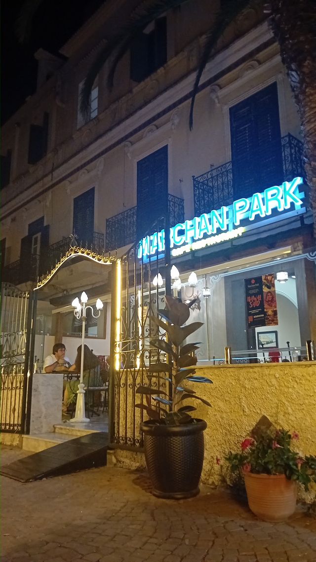 L'entrée de Marchan Park la nuit : la grille en fer forgé, les lampes et les plantes devant l'enseigne néon verte