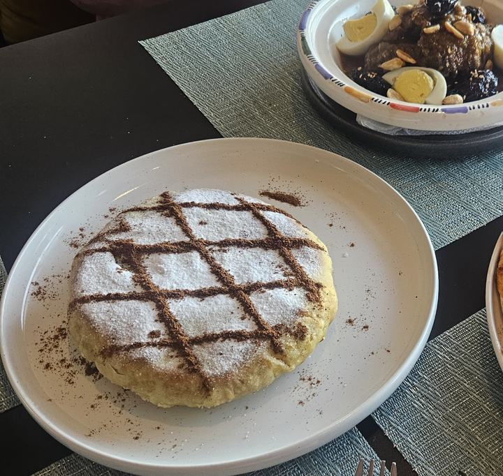 Pastilla dorée saupoudrée de sucre glace et de cannelle, avec un tajine de viande aux pruneaux, amandes et œufs en arrière-plan
