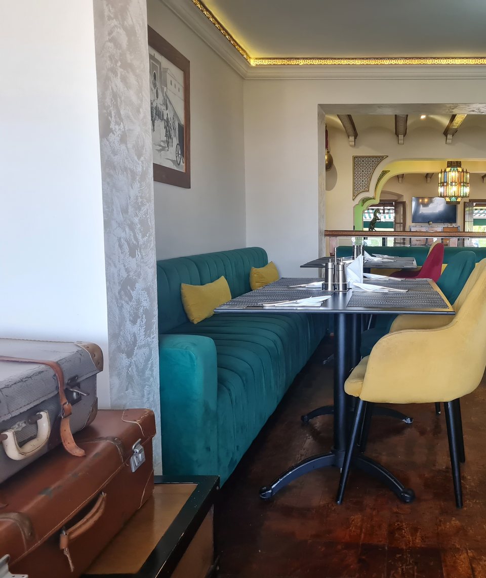 La salle : banquette en velours bleu-canard, chaises moutarde, valises anciennes empilées en décor, sous les arches et une lanterne